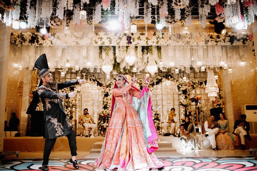 Bride and groom performing a dance number on stage as guests watch, beneath an elaborate hanging chandelier installation, Zara & Shuaib's wedding at Garrison Halls, Lahore