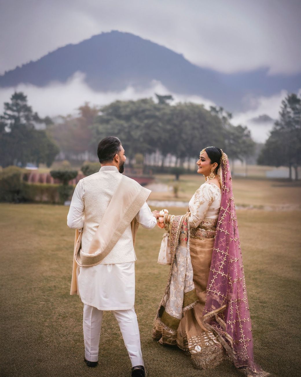 Bride and groom walking hand in hand on a misty hillside, glancing back over their shoulders, mountains in the background