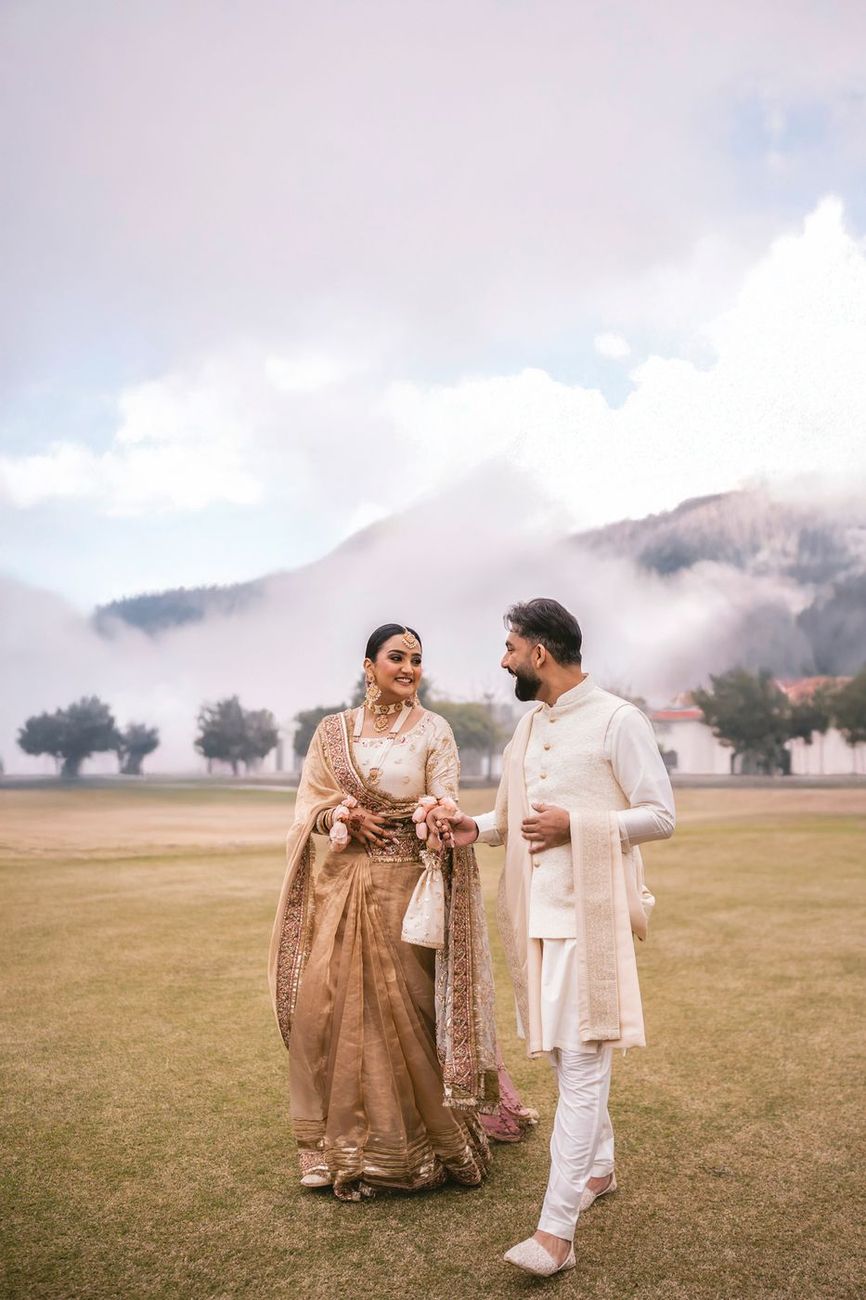Wide shot of the couple walking across the misty field, mountains and a building in the distance