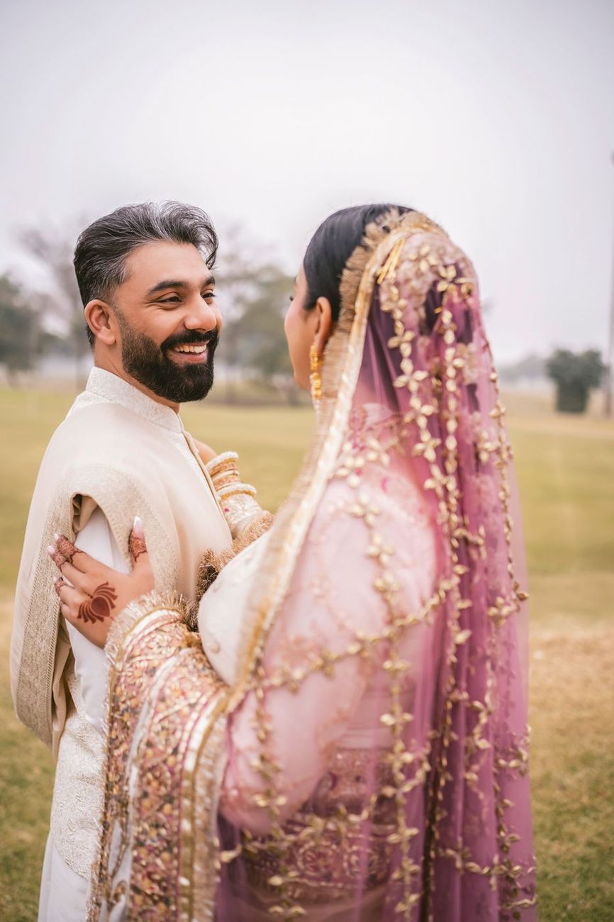 Reverse angle of the groom laughing as he looks at the bride, her mauve embroidered dupatta visible