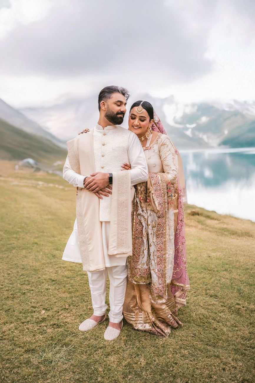 Couple embracing on a grassy hillside above a turquoise glacial lake, snow-capped mountains behind