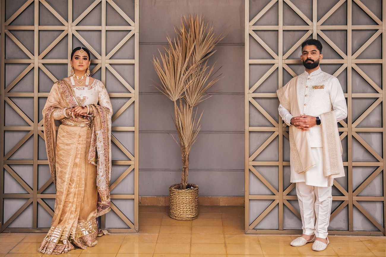 Bride and groom standing apart on either side of a potted palm against a geometric latticed wall