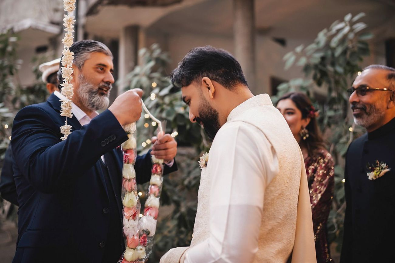 Groom bowing his head while an older relative places a floral garland around his neck