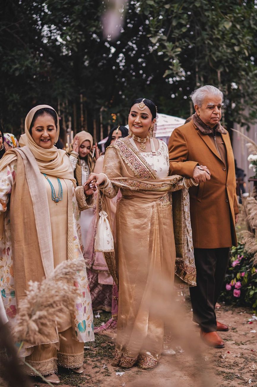 Bride's entrance, holding hands with both parents as they walk through the garden venue