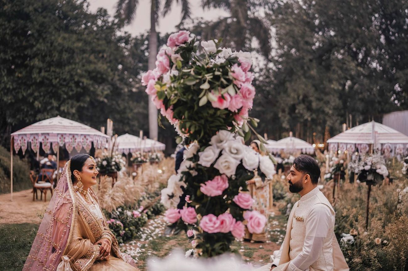 Wide nikah ceremony shot, bride and groom seated facing each other, separated by a tall floral arrangement