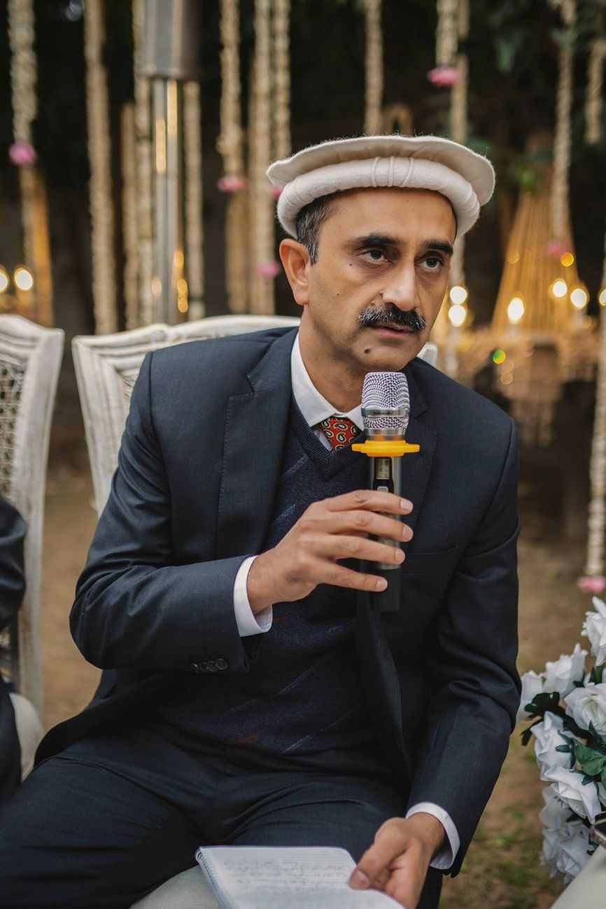 Man in a traditional cap reading from notes into a microphone during the ceremony