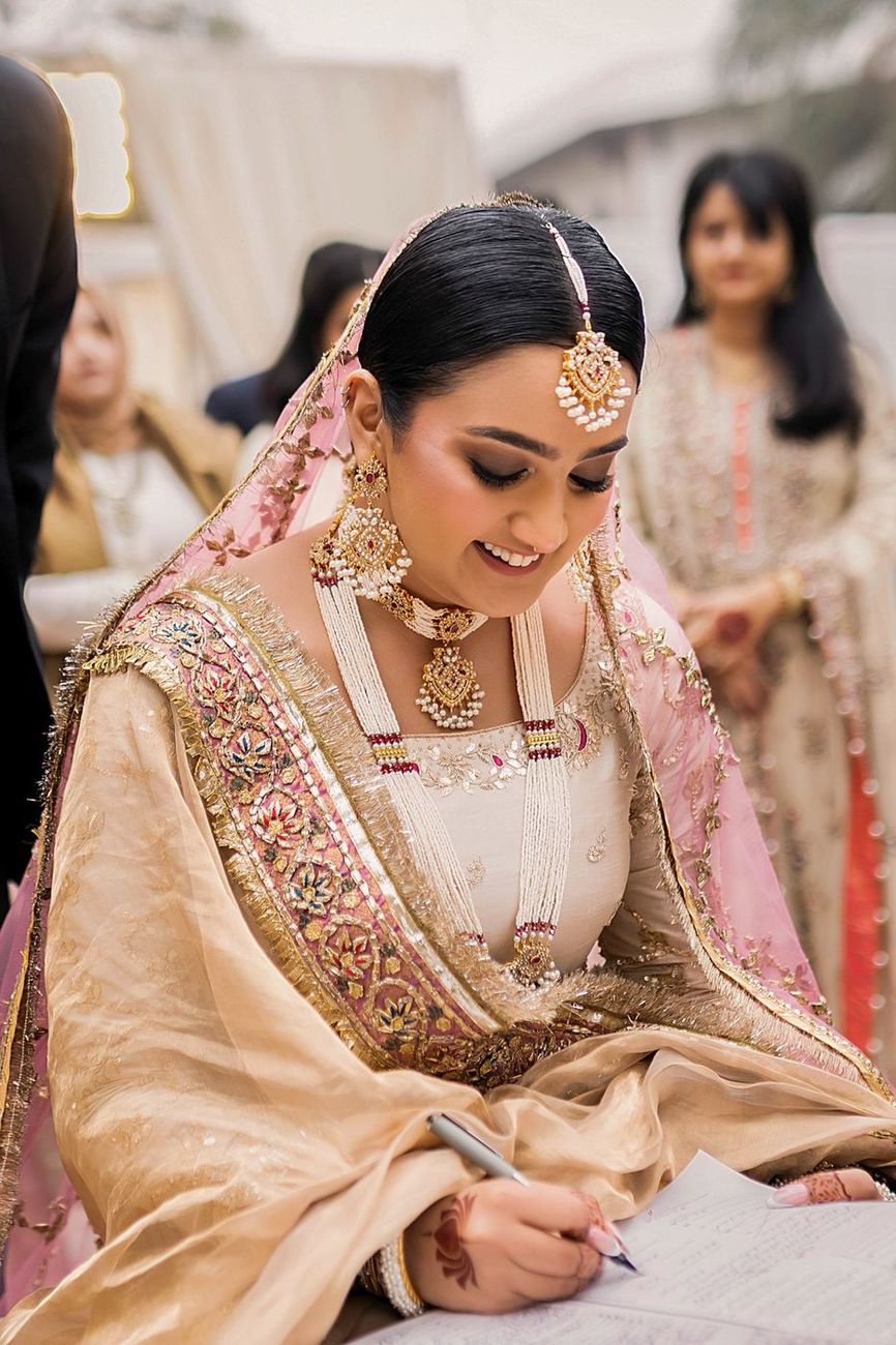 Bride signing the nikah documents, smiling, full bridal jewelry visible