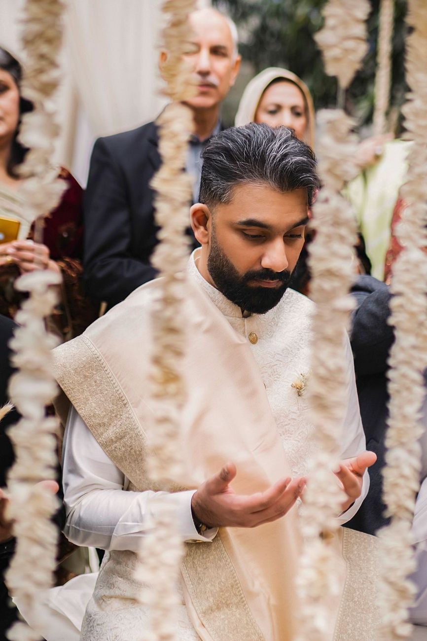 Groom in a dua pose, hands cupped in prayer, framed through hanging garlands