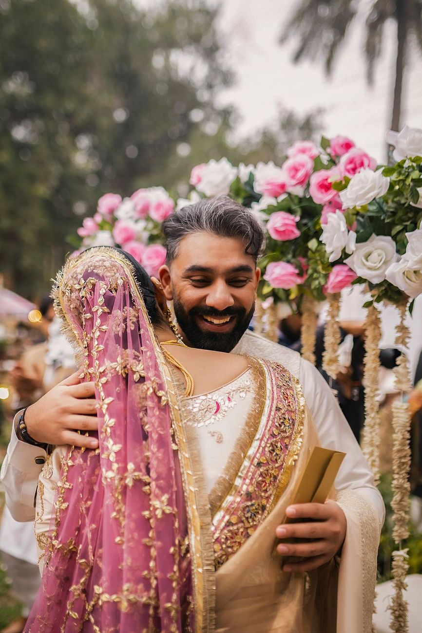 Groom embracing the bride while holding an envelope, pink and white roses behind them