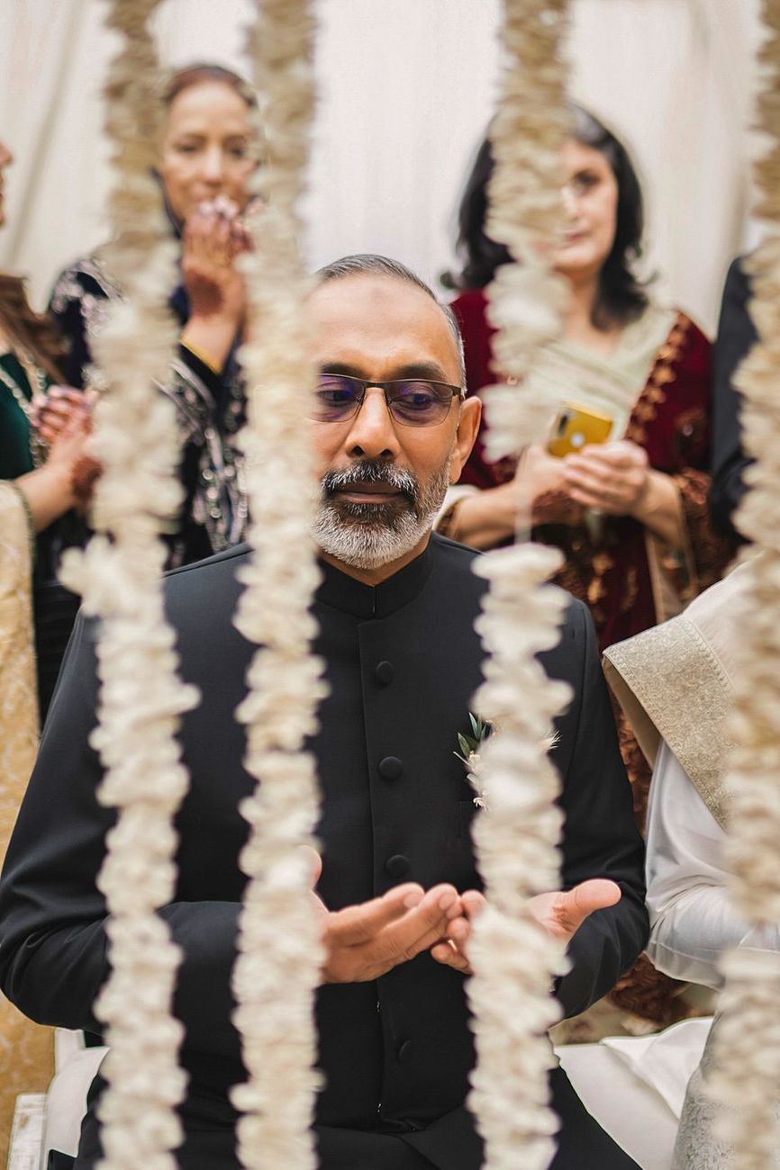 Groom's father caught mid-speech, framed through hanging garland strands