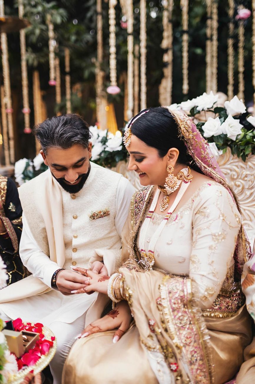 Groom placing a ring on the bride's finger, both seated on the ornate floral bench