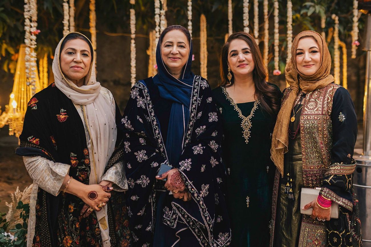 Four women posed together in dark jewel-toned outfits, evening lighting and garlands behind
