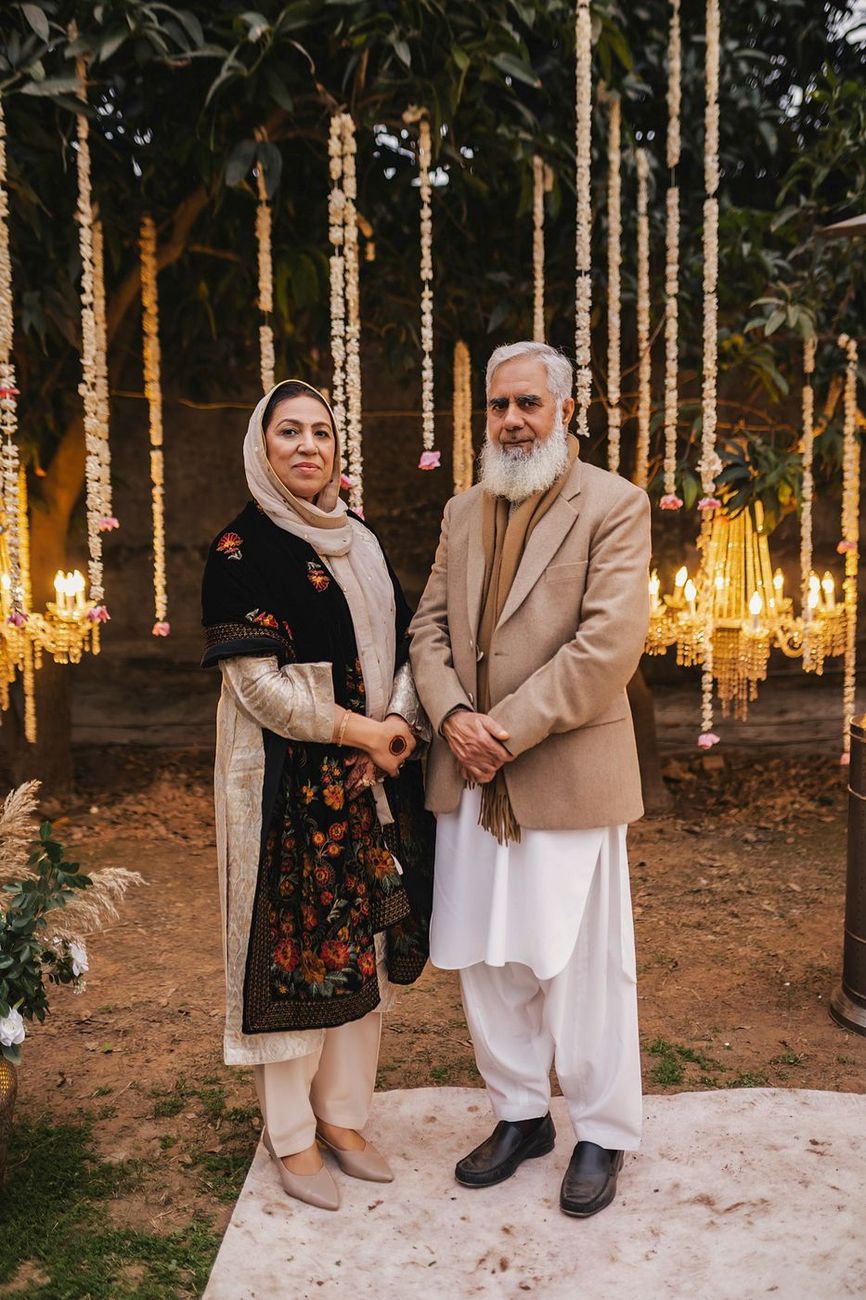 Older couple, the man with a long white beard, posed near the lit chandeliers and hanging garlands