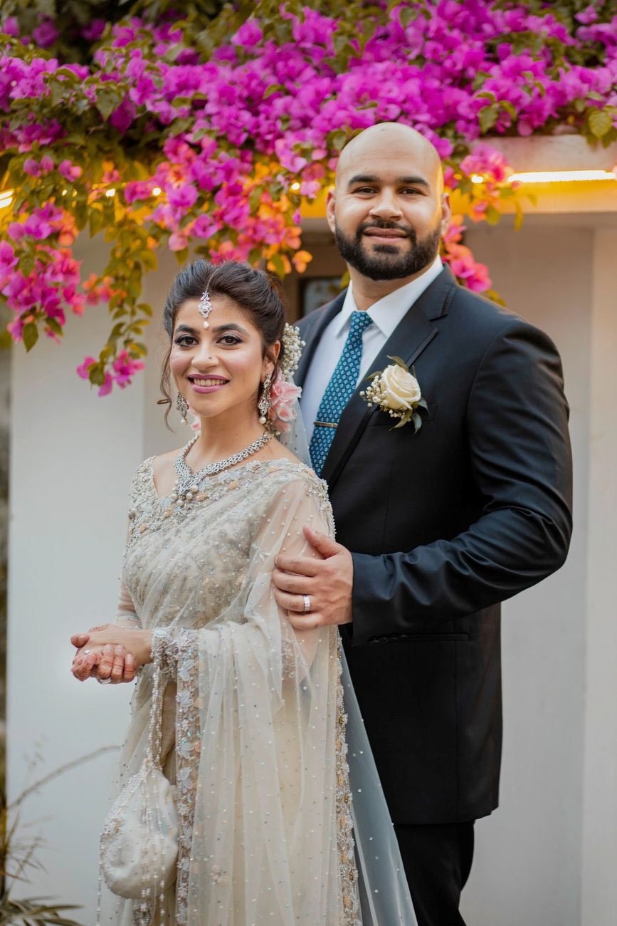 Bride in an ivory embellished gown and groom in a navy suit smiling under a bougainvillea-draped wall, Mashal & Danial's wedding at a private residence in Lahore