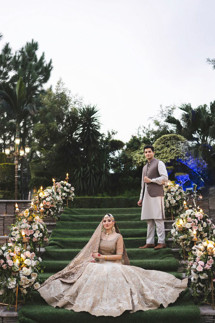 Bride and groom walking down a grass-covered staircase lined with floral arrangements, holding hands, Nayha & Rafay's wedding in Islamabad