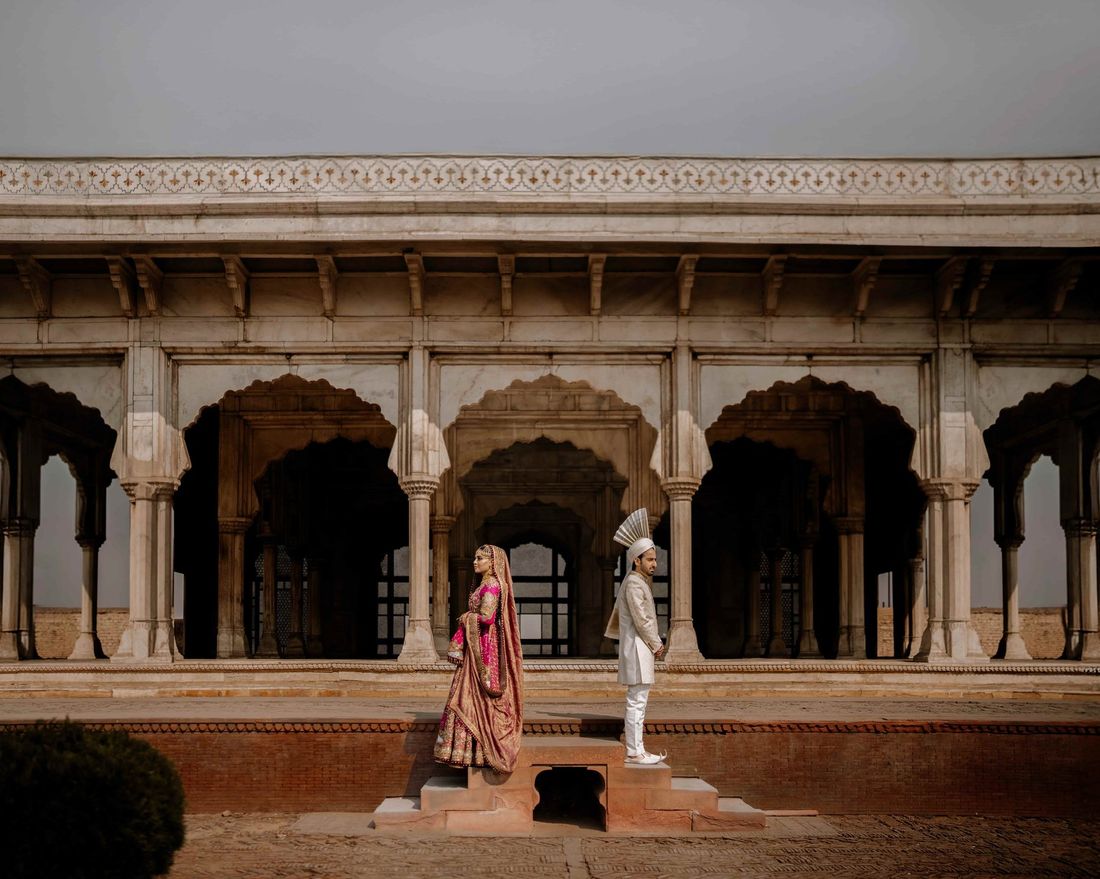 Bride and groom in traditional wedding attire standing before a grand marble colonnade with arched alcoves