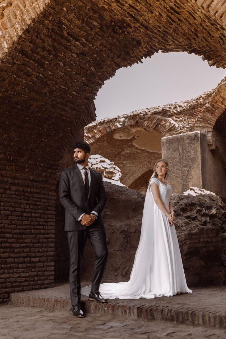Bride in a flowing white gown and groom in a dark suit standing within ancient brick ruins