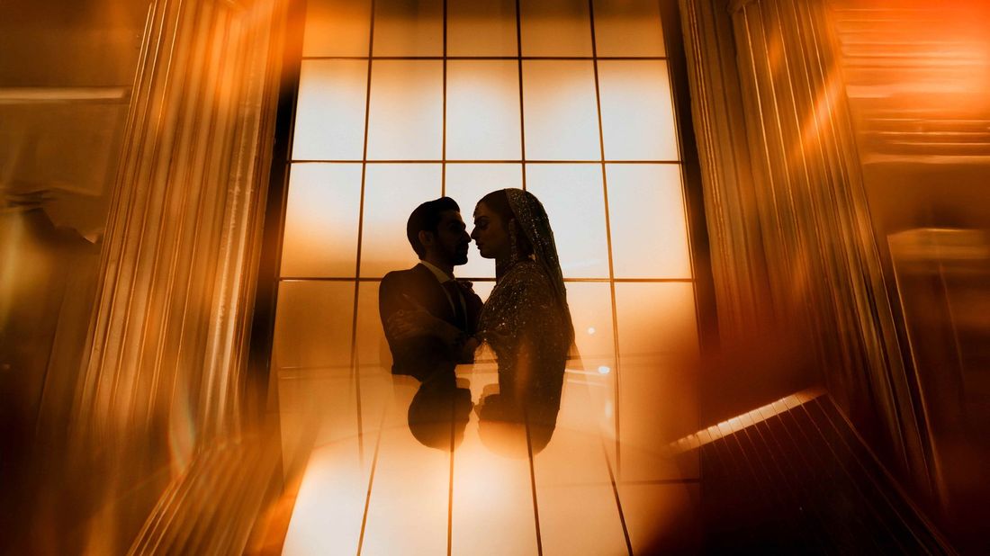 Silhouette of bride and groom standing close together against a glowing backlit window, reflected on the floor below