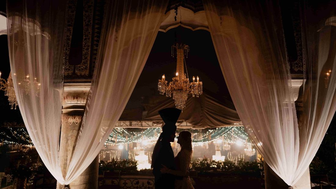 Silhouette of bride and groom beneath a chandelier and draped curtains, overlooking a festively lit reception venue at night