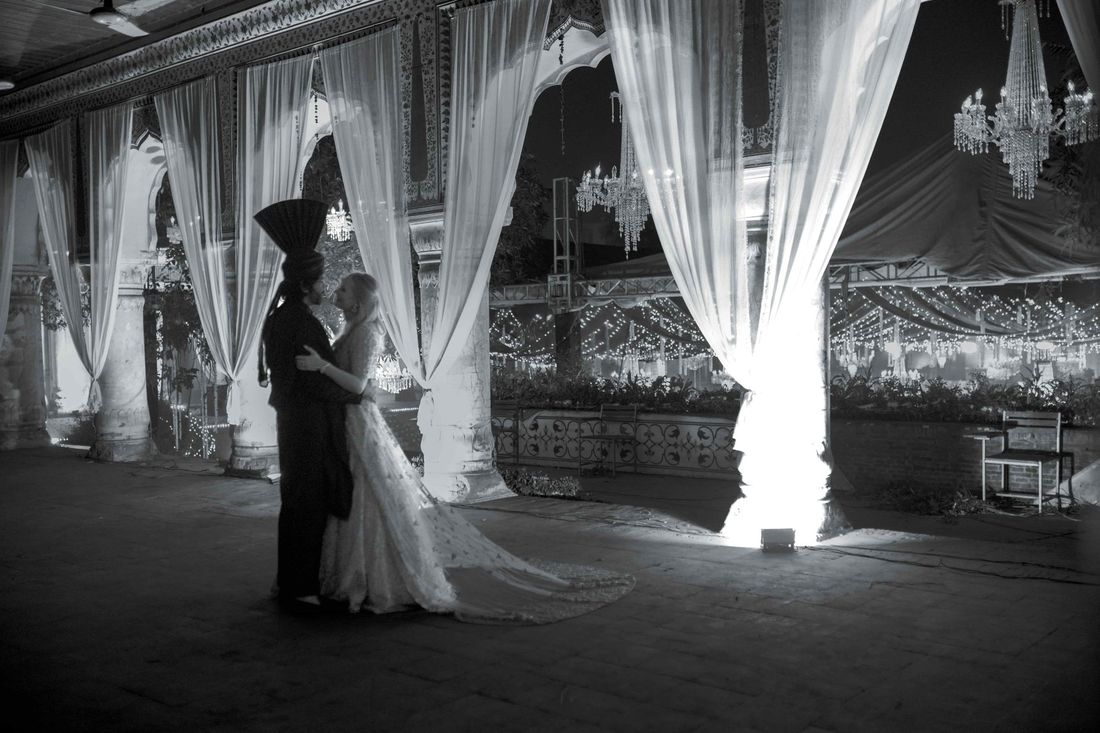 Black-and-white photo of bride and groom embracing beneath a draped colonnade with chandeliers, overlooking a string-lit garden reception
