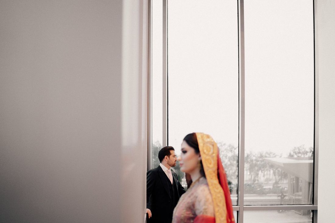 Bride in a red and gold dupatta in close-up foreground, groom in a formal black suit watching from behind a glass wall