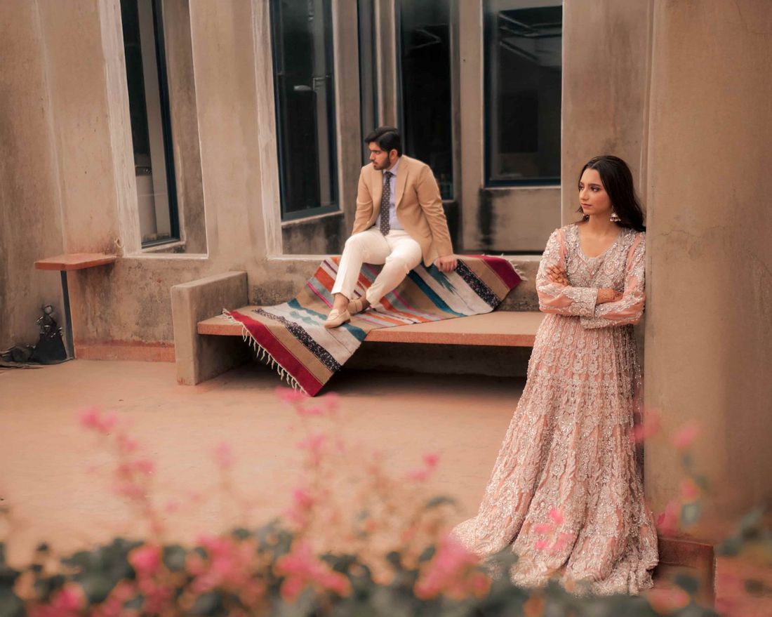 Bride in a blush embellished gown leaning against a terracotta pillar, groom seated on a rug-draped bench in the background, with bougainvillea blooms in the foreground