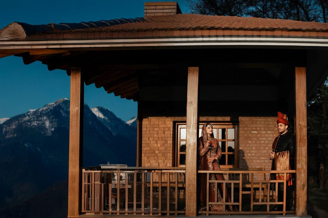 Bride and groom standing on a wooden mountain lodge porch with snow-capped peaks in the background at dusk