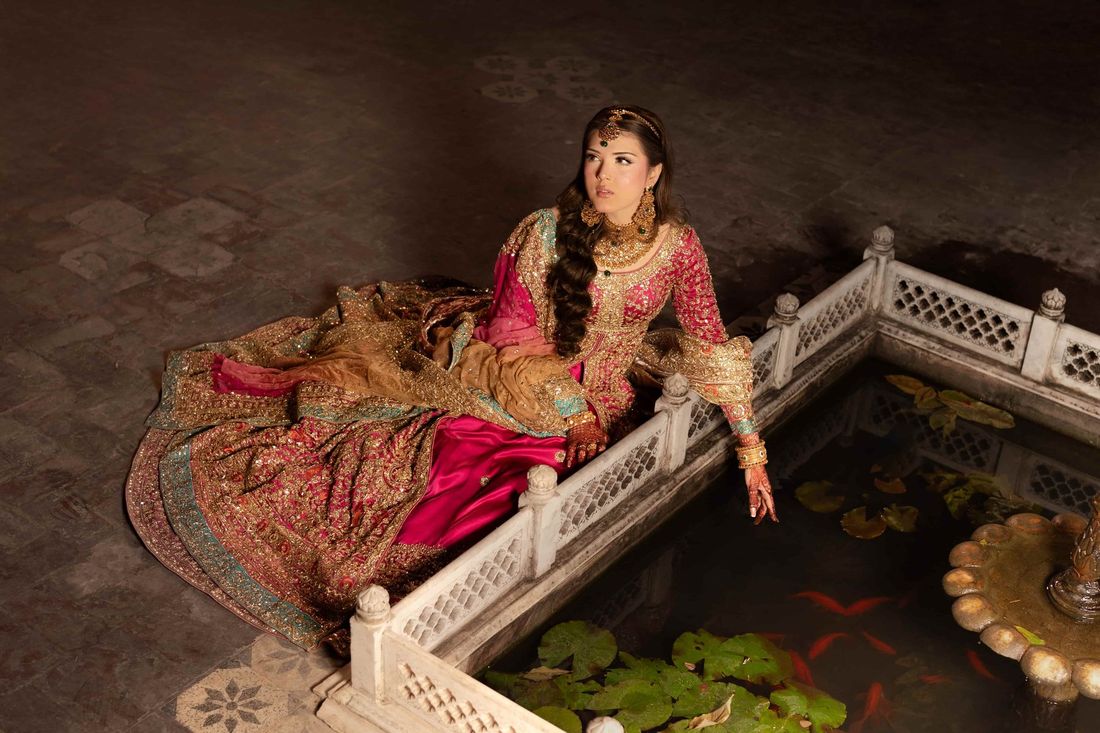Bride in an ornate fuchsia and gold bridal lehenga reclining beside a marble fountain with koi fish