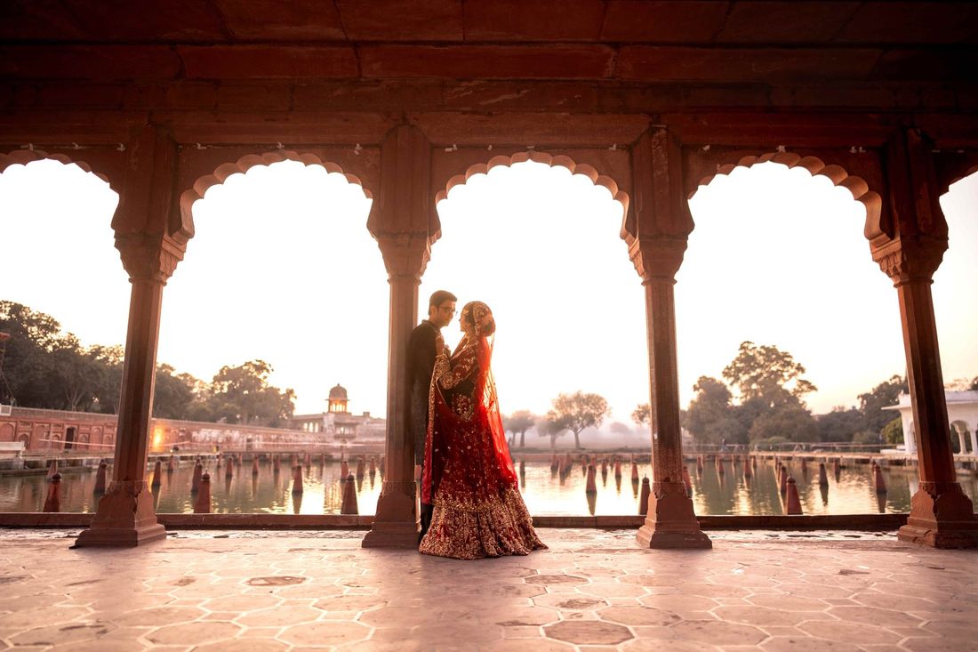 Bride and groom embracing beneath red sandstone scalloped archways overlooking a tranquil lake at golden hour