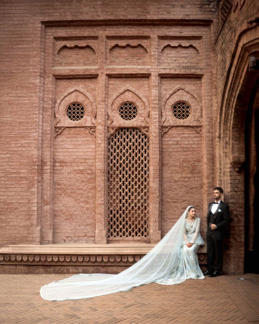 Bride in a pale blue gown with a long flowing train, groom in a black tuxedo, standing before an ornately carved brick facade with lattice windows