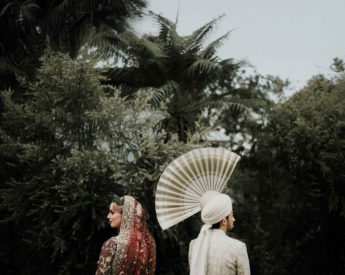 Bride and groom in profile against lush greenery, groom wearing a white turban with a tall fan-shaped kalgi