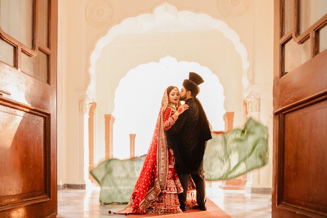 Groom kissing the bride's forehead in an intimate moment, framed by grand wooden doors and a Mughal archway