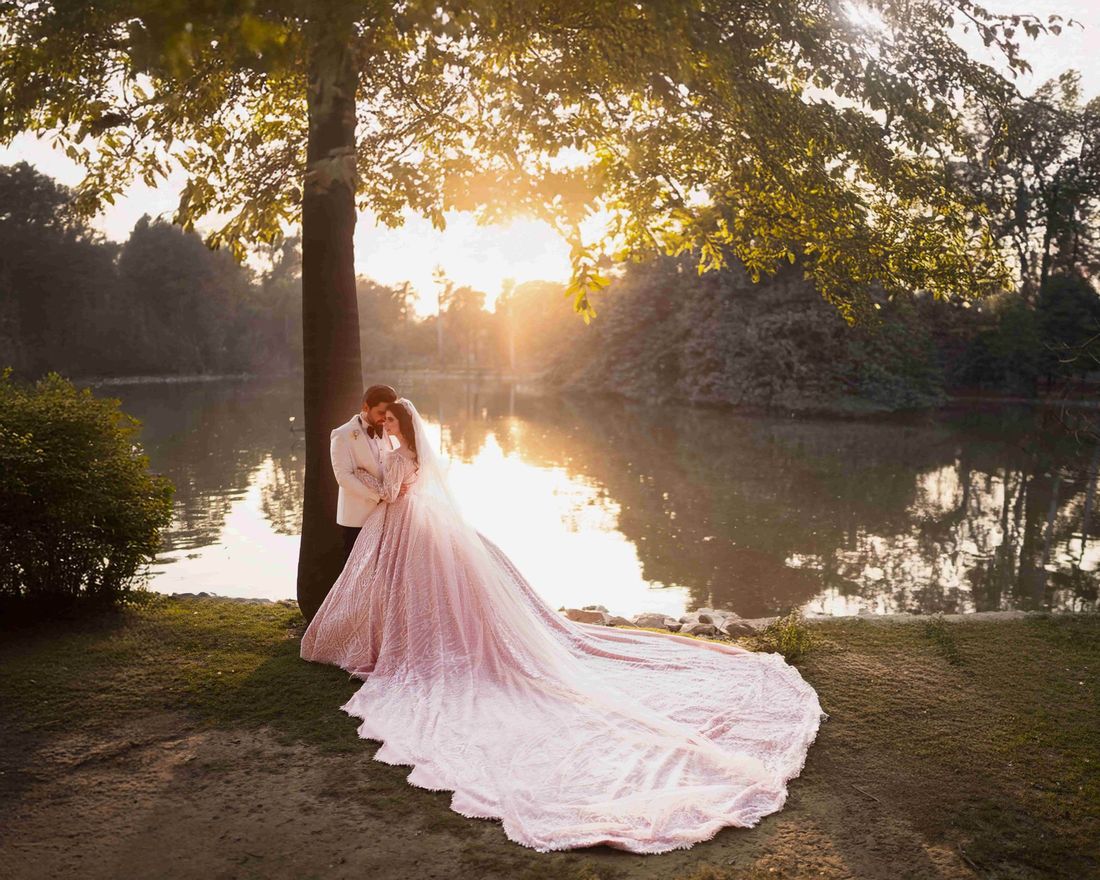 Bride in a blush pink ballgown with a sweeping train and groom in a white tuxedo jacket, embracing beside a lake at sunset
