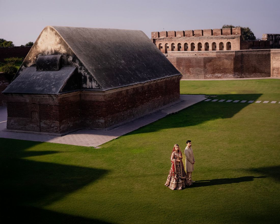 Bride and groom standing on a vast lawn beside a historic curved-roof fort building, viewed from above
