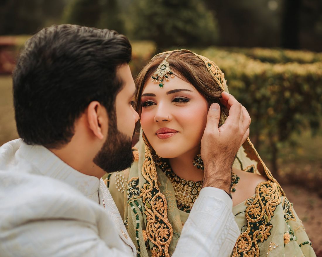 Close-up of groom gently touching the bride's veil as she smiles, bride wearing a green and gold embroidered dupatta with emerald jewelry