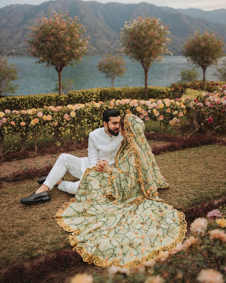 Bride and groom seated together beside a flower garden with a lake and mountains behind them, bride's green and gold lehenga spread across the grass