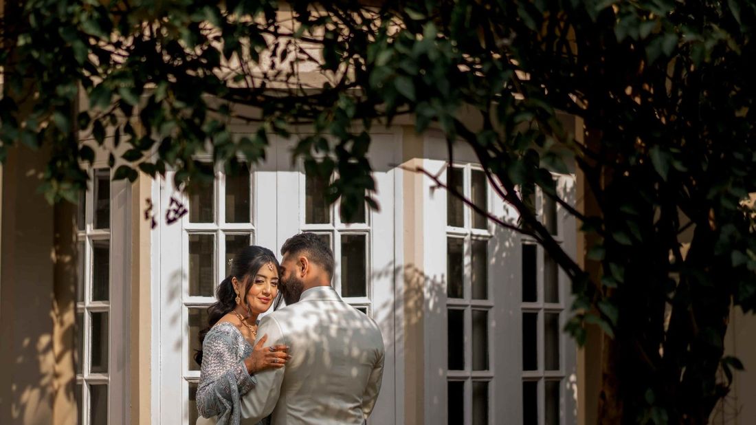 Bride and groom embracing in front of white colonial-style French doors, framed by dappled tree shadows