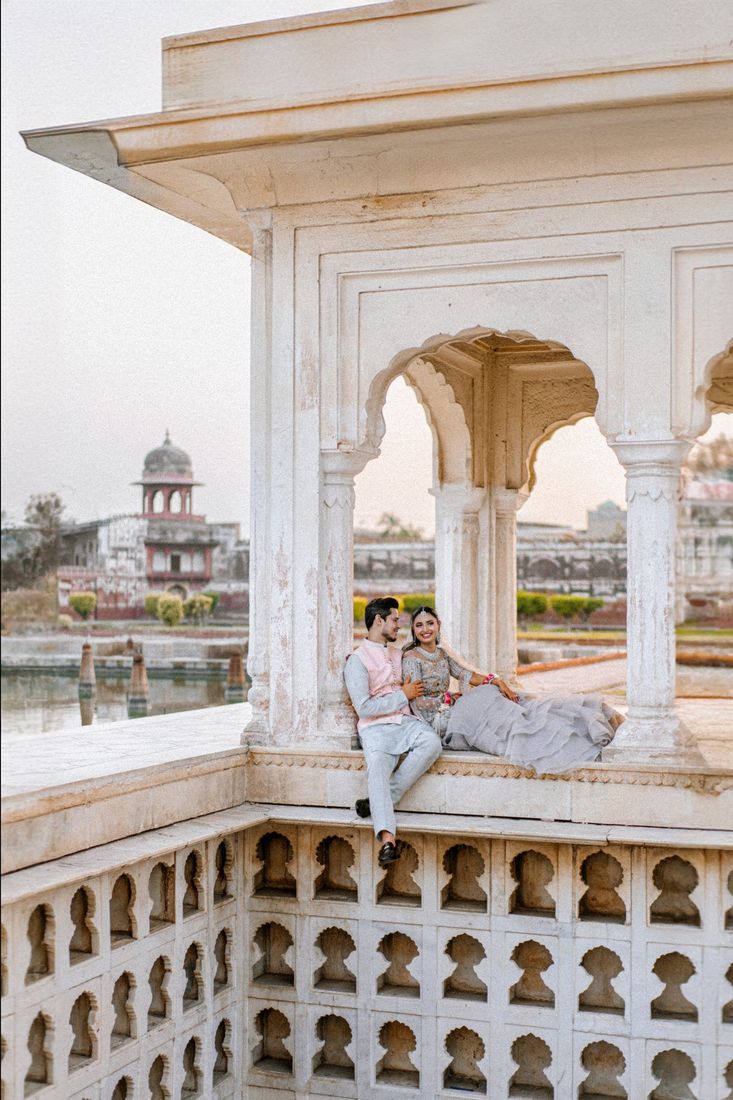 Bride and groom seated together within a white marble pavilion with scalloped arches, overlooking a Mughal garden and lake