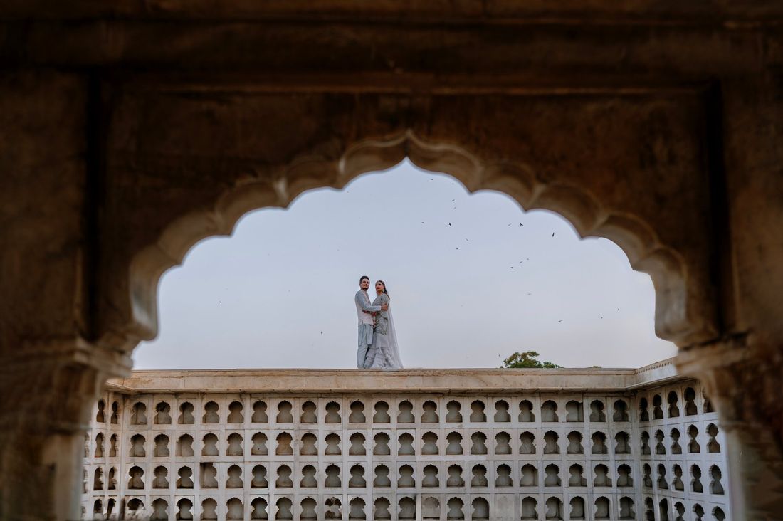 Bride and groom standing atop a honeycomb-patterned wall, framed within a grand scalloped archway with birds in flight