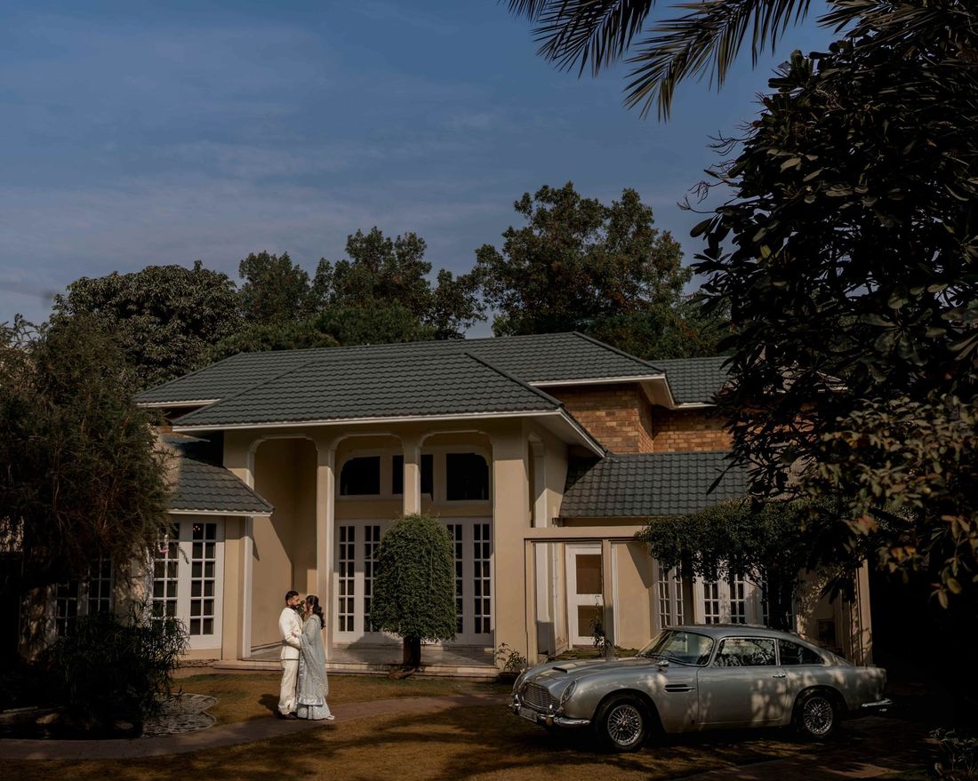 Bride and groom standing together in front of a grand residence, a vintage silver sports car parked beside them