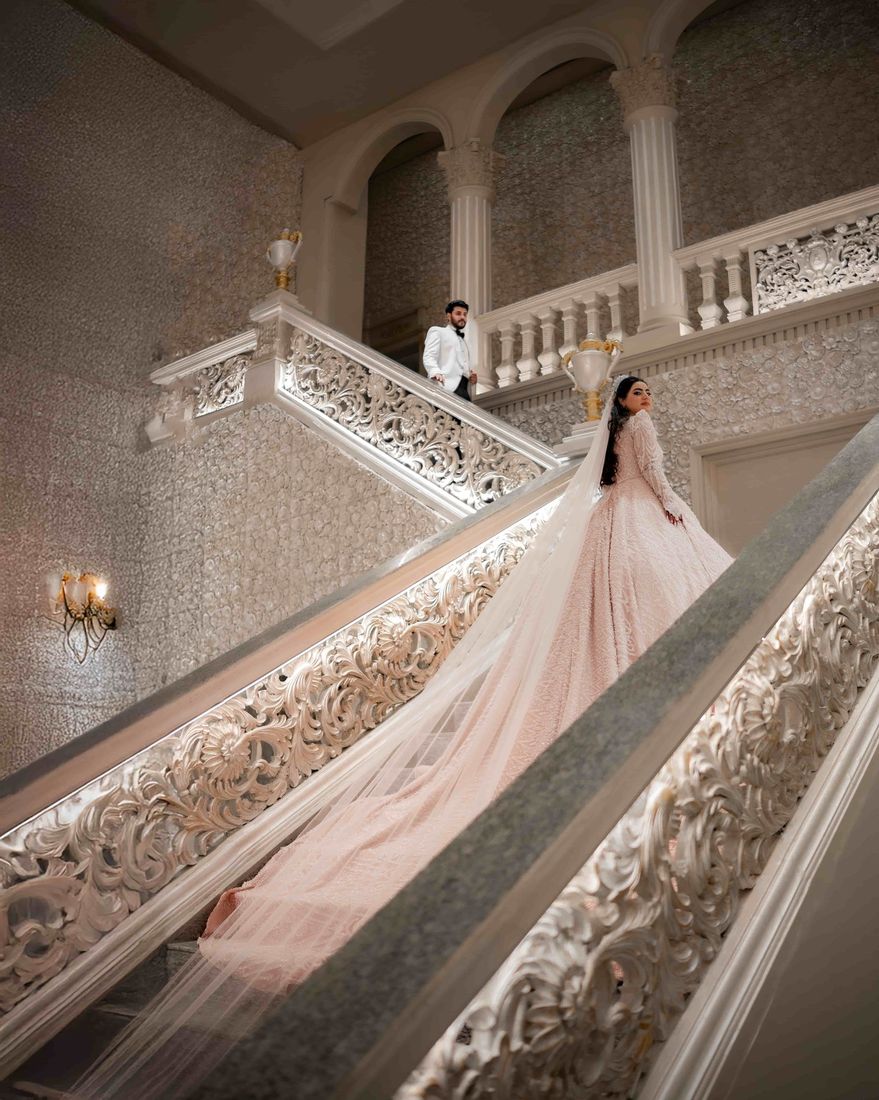 Bride glancing over her shoulder on the grand staircase, her pink gown train flowing down the ornate steps