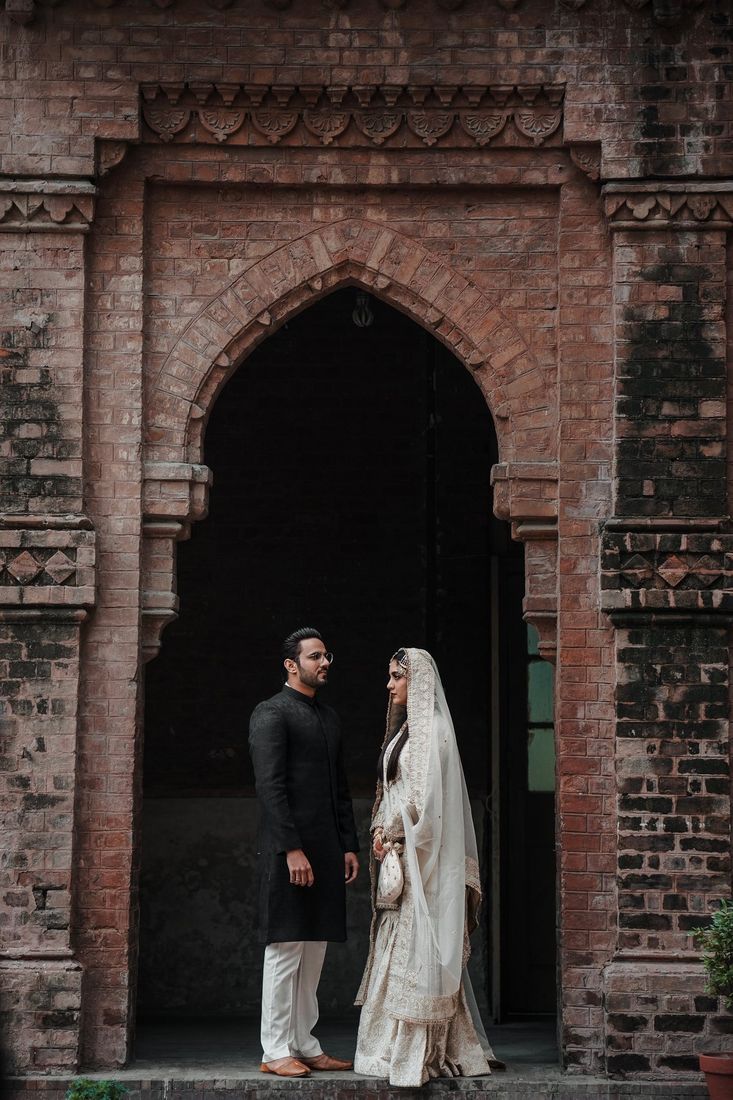 Bride in an ivory embroidered outfit and groom in a black kurta standing within a tall pointed brick archway