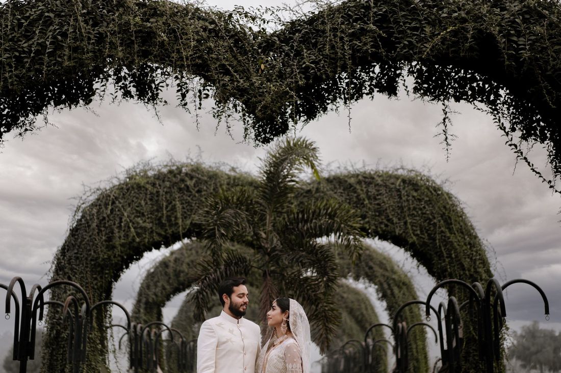 Bride and groom in ivory outfits standing beneath a row of vine-covered archway trellises under an overcast sky