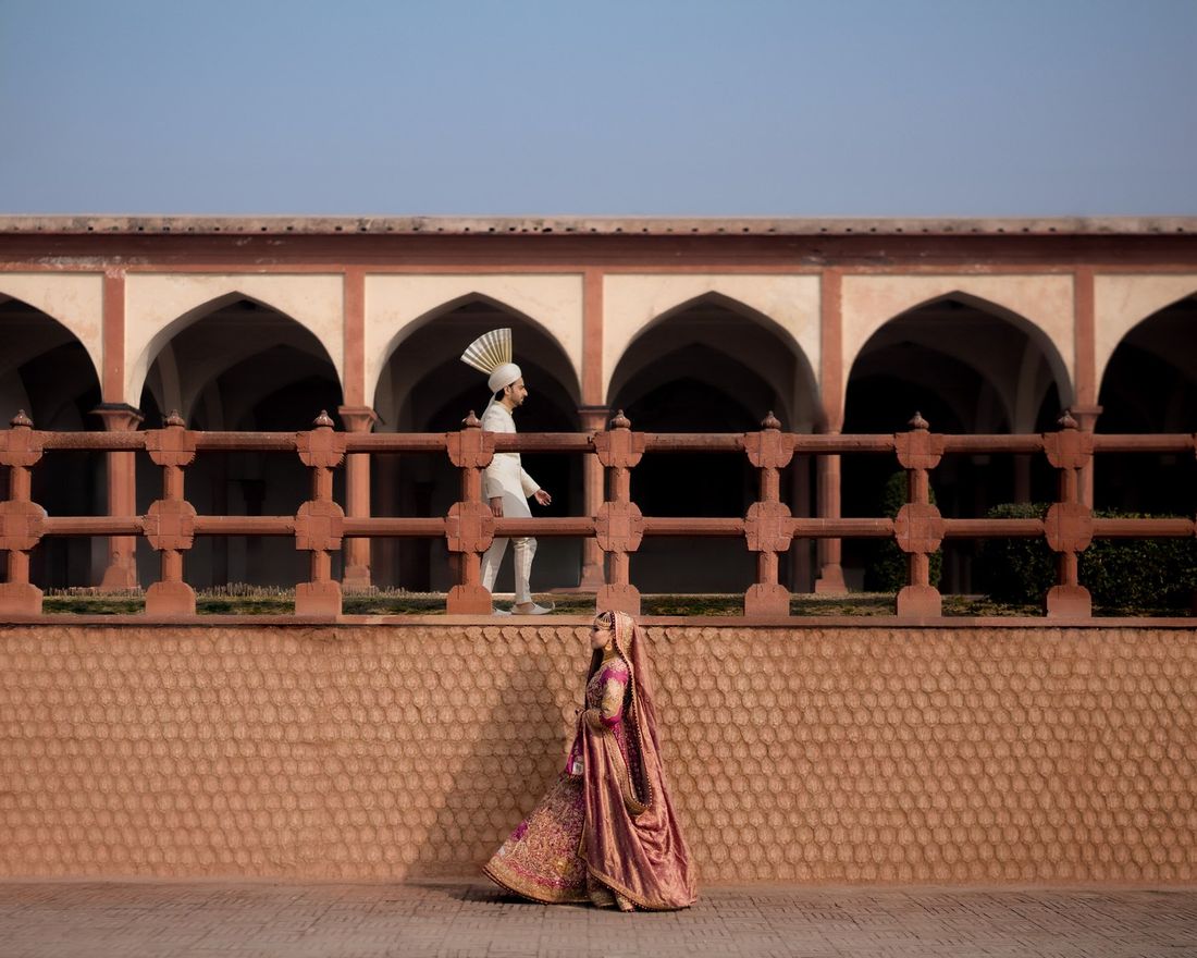 Groom walking along an upper arcade balustrade while the bride stands below against a honeycomb-patterned wall