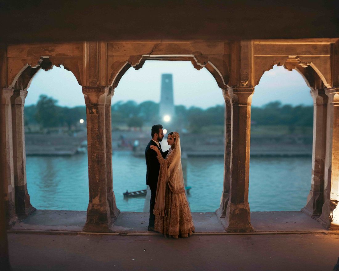Bride and groom embracing beneath red sandstone arches overlooking a river at dusk, a small boat and tower visible in the distance