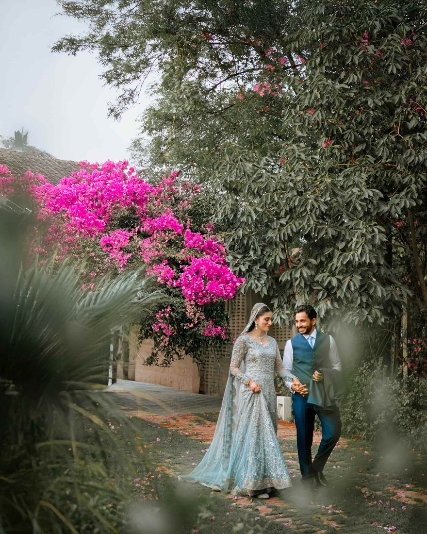 Bride and groom walking together along a brick garden path beneath a vivid pink bougainvillea bush
