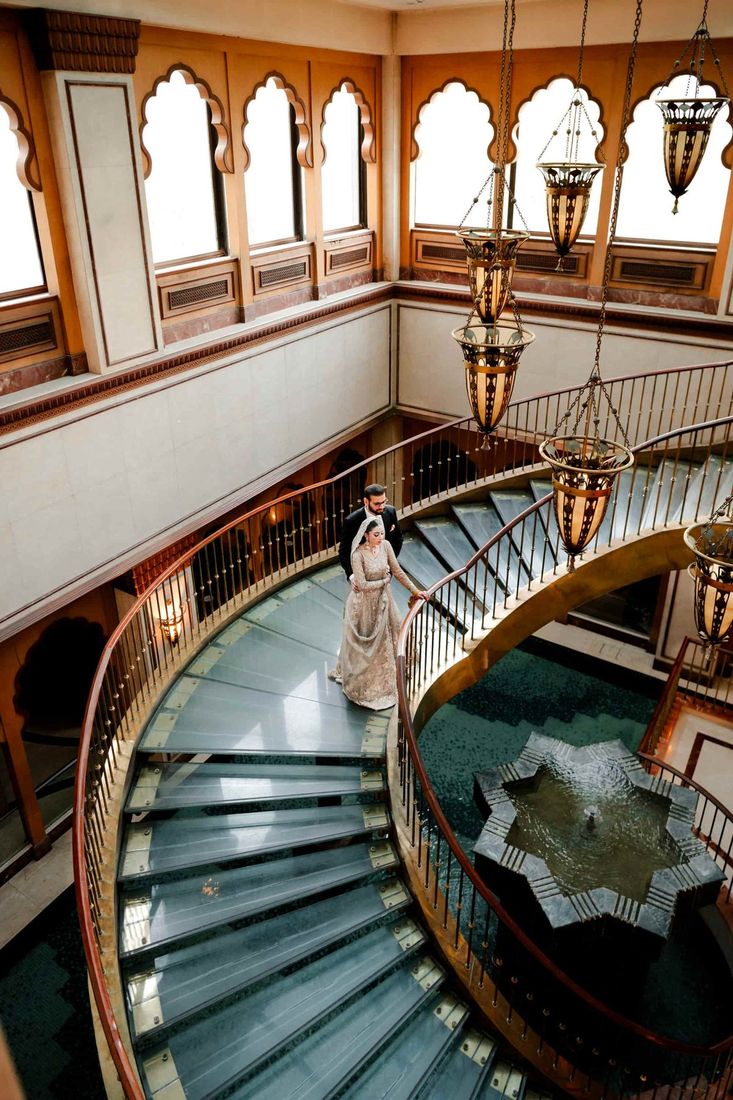 Bride and groom descending a curved staircase above a star-shaped fountain, beneath ornate hanging lanterns and Moorish-arched windows