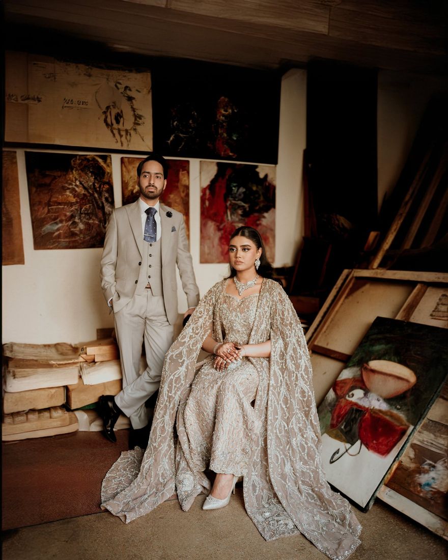 Bride in an embellished cape-style gown seated beside the groom in a beige three-piece suit, set in an artist's studio surrounded by paintings