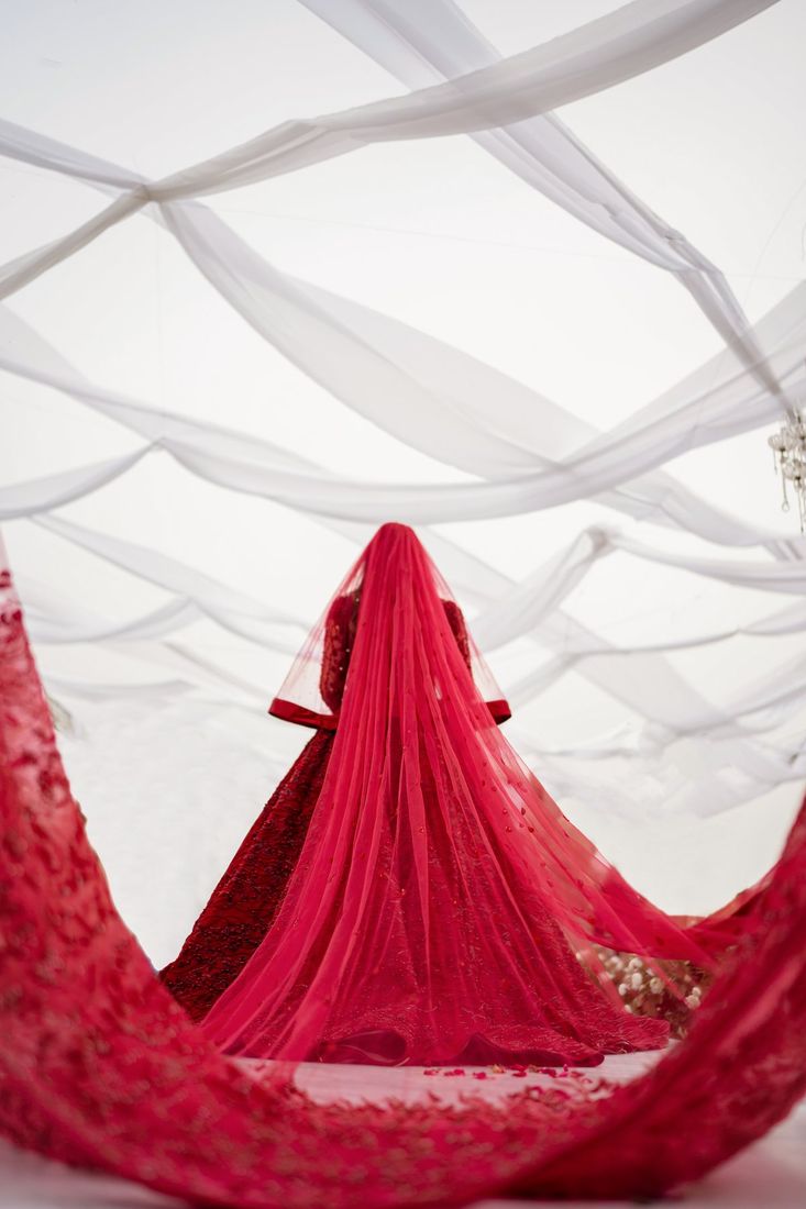 Bride from behind with a deep red veil cascading down, dress fanned out in a wide circle, framed by white draped tulle streamers