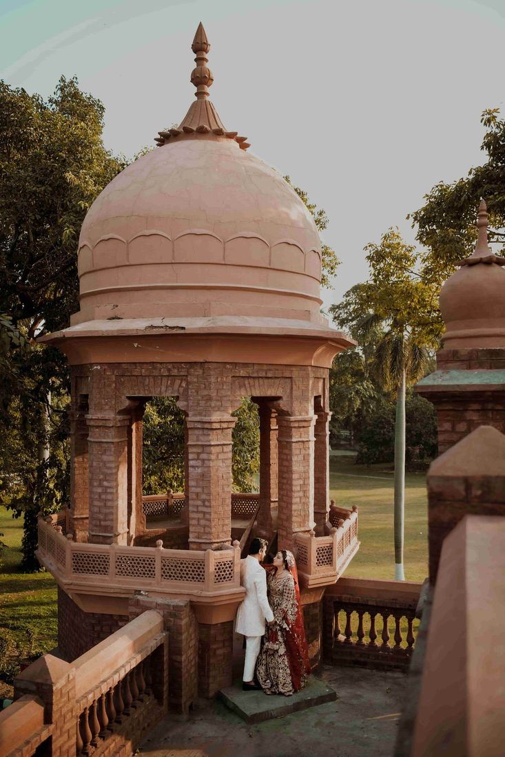 Bride and groom standing close together at the base of a domed heritage pavilion, surrounded by lawns and trees